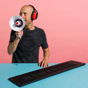 Man in red headphones speaking into a megaphone behind a keyboard controller against a pink background