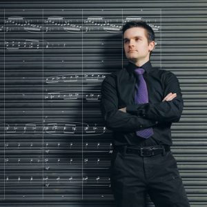 JJ Berthume standing with arms crossed in a black shirt and purple tie in front of sheet music